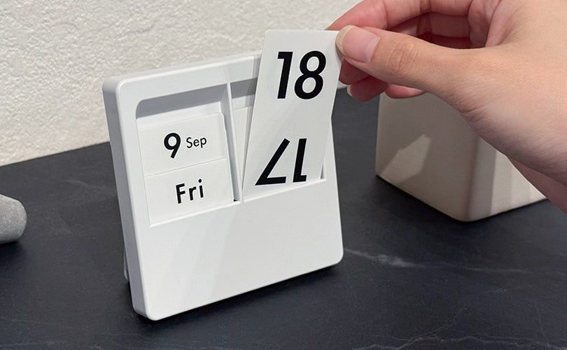 Hand changing a date panel on a white Carl Panel Calendar on a desk