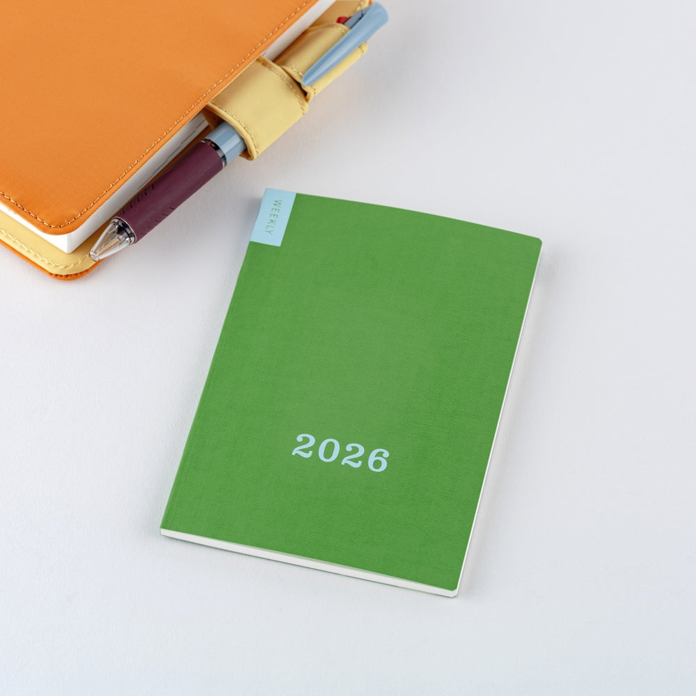 Angled view of green Hobonichi Weekly 2026 calendar next to orange notebook