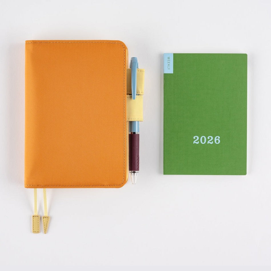 Green Hobonichi Weekly 2026 pocket calendar flat lay with orange notebook