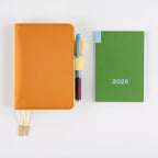 Green Hobonichi Weekly 2026 pocket calendar flat lay with orange notebook
