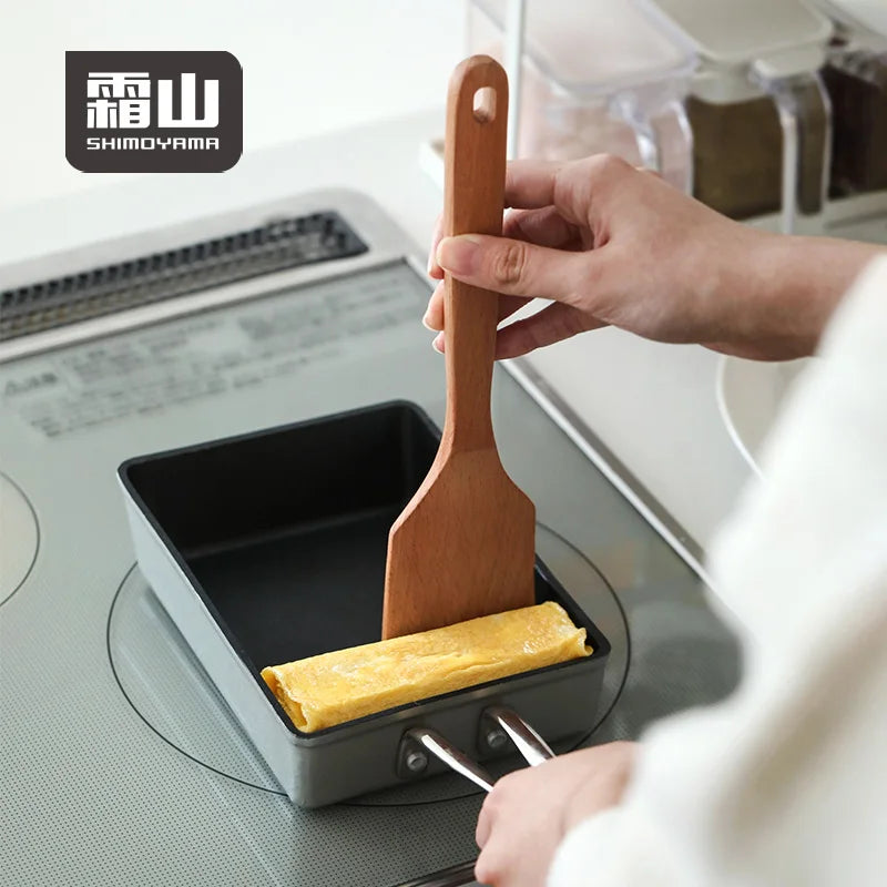 Pressing square wooden spatula against tamagoyaki in black pan