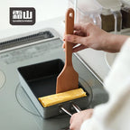 Pressing square wooden spatula against tamagoyaki in black pan
