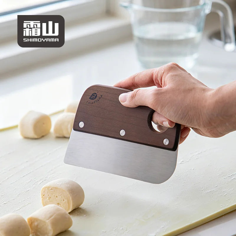 Hand holding Shimoyama dough scraper by circular hole in dark wood handle above cut dough pieces