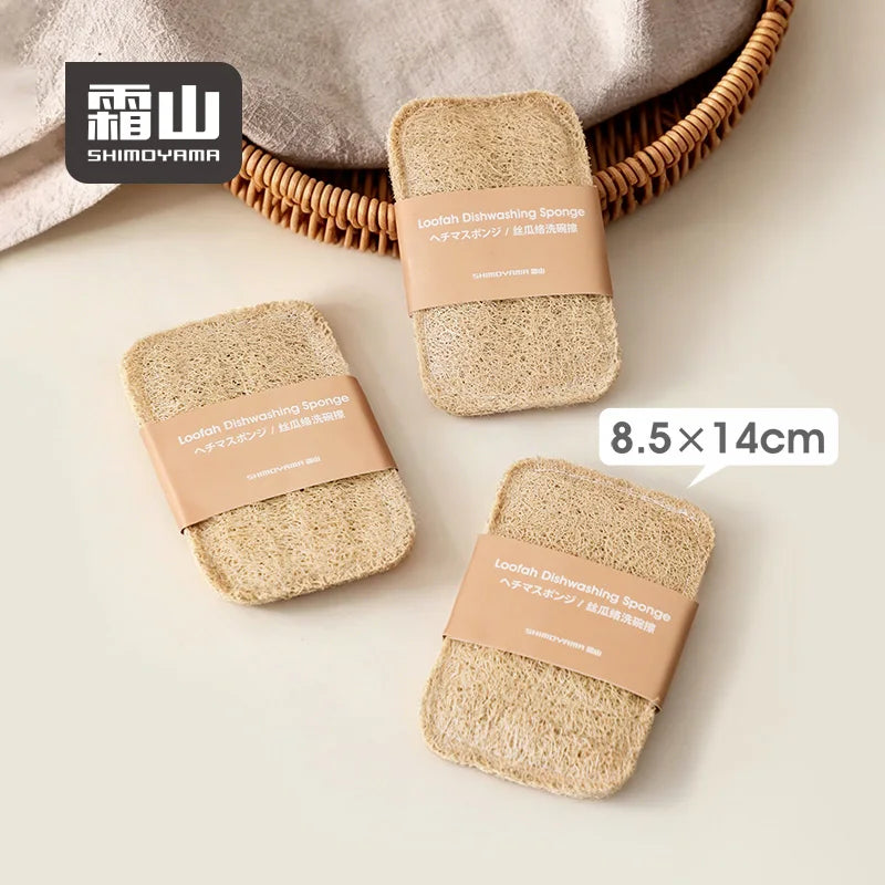 Three packaged loofah dish wipes next to linen cloth and wicker basket