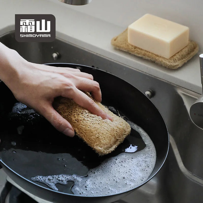 Hand using beige loofah scrubber to clean soapy black pan in sink