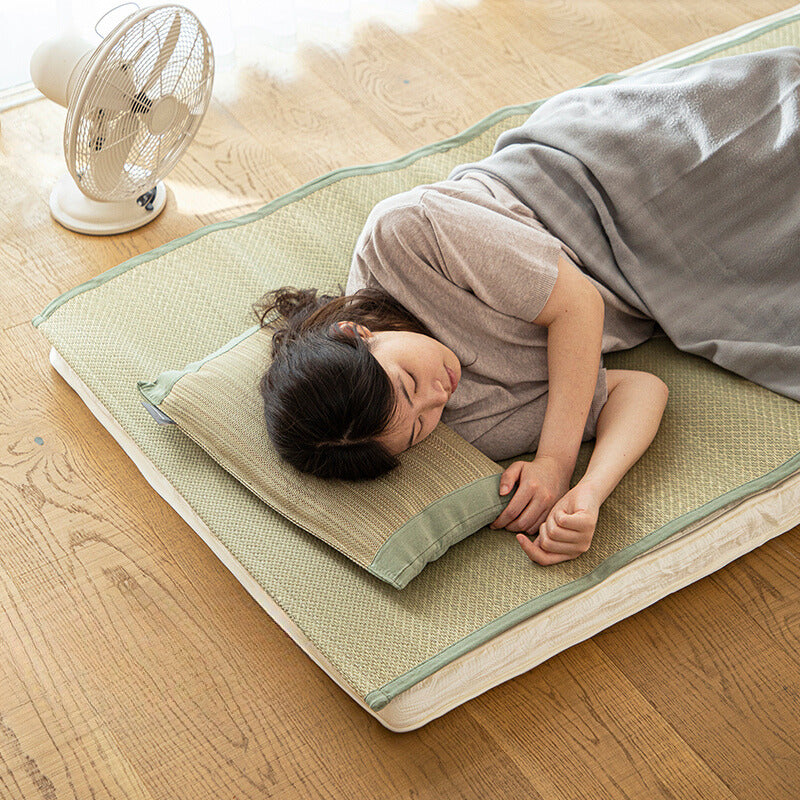 Person using olive green trimmed rush straw pillow on wooden floor with fan