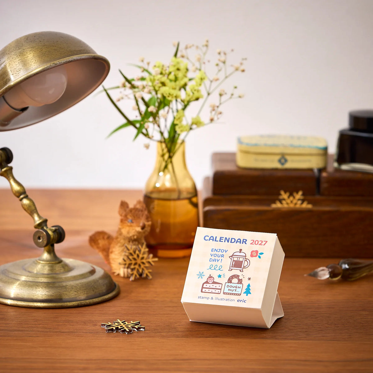 eric Colour Daily Calendar 2027 on a wooden desk beside a lamp and flowers