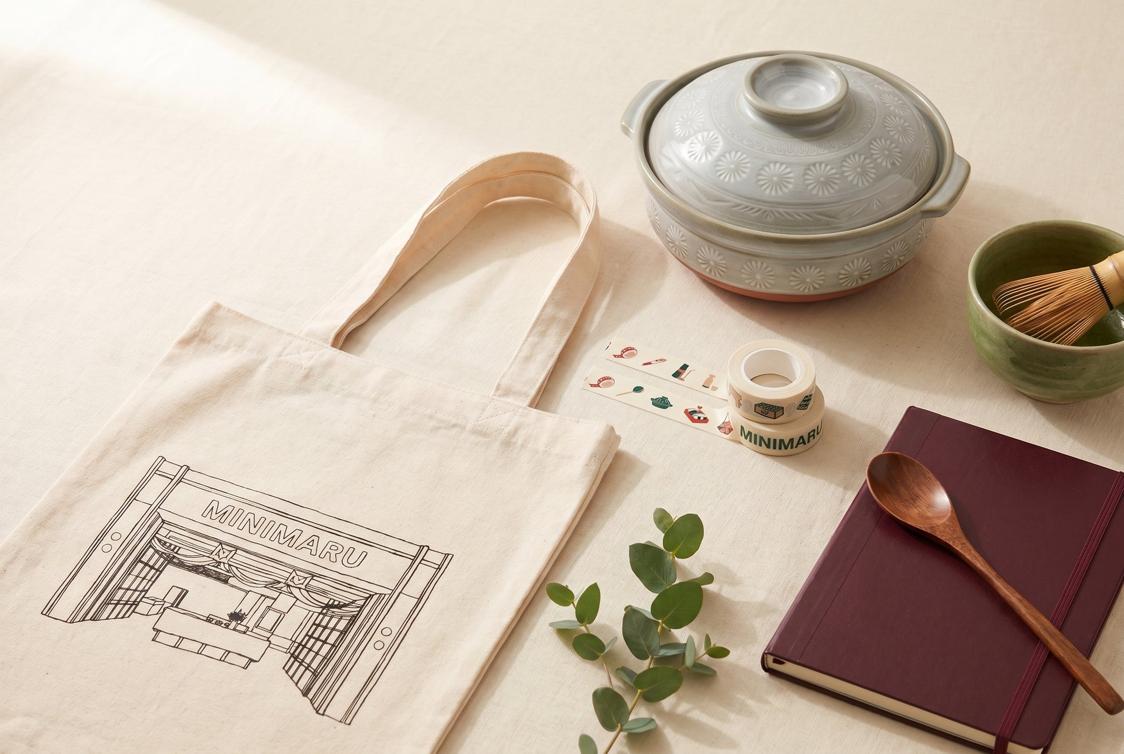 Minimaru anniversary flatlay — donabe pot, matcha bowl, washi tape, Hobonichi notebook and canvas tote bag on a cream linen surface