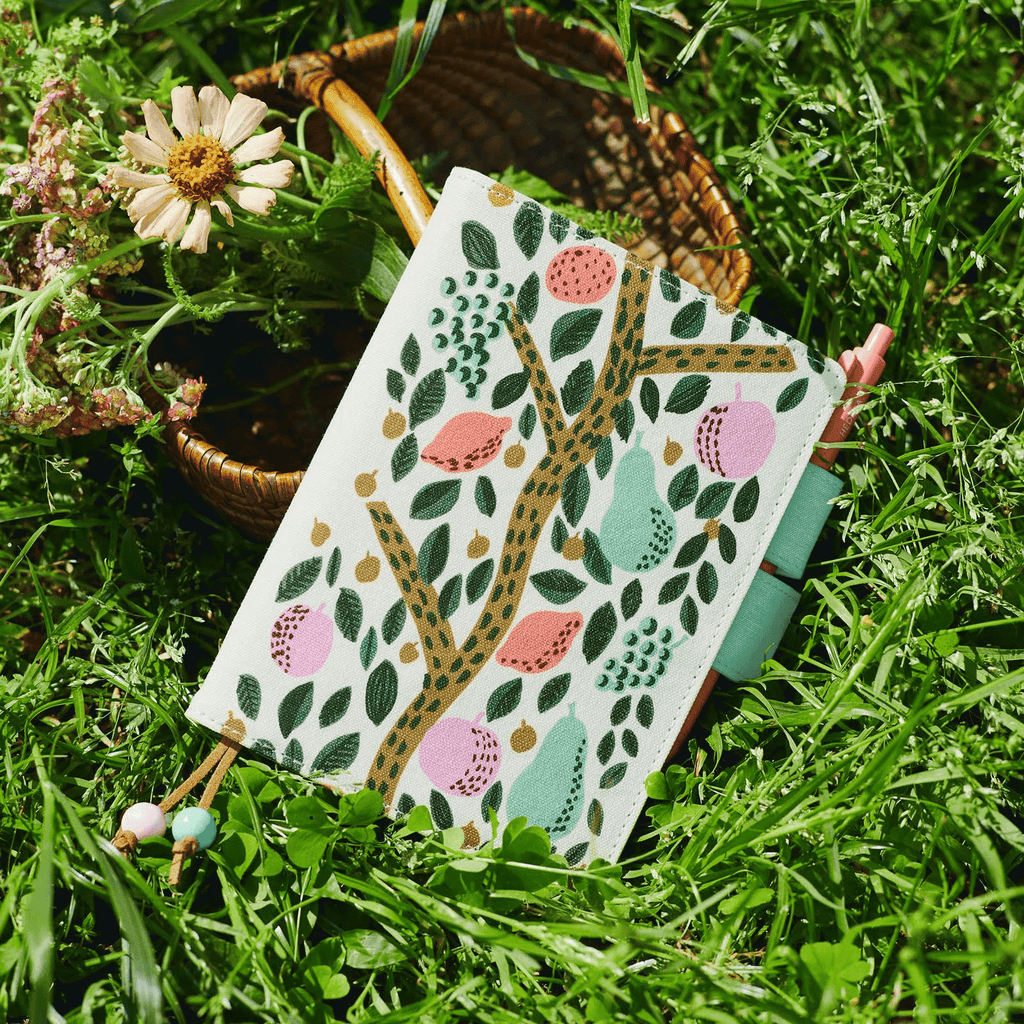 Hobonichi Techo A6 planner with botanical print resting in grass beside wildflowers and pen tucked in elastic band
