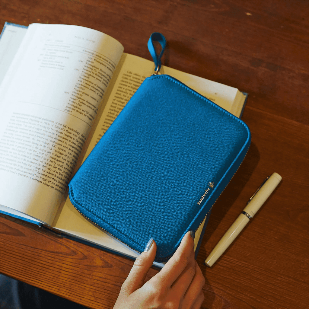 Hand holding blue Mare Hobonichi planner with wrist strap on wooden desk with pen