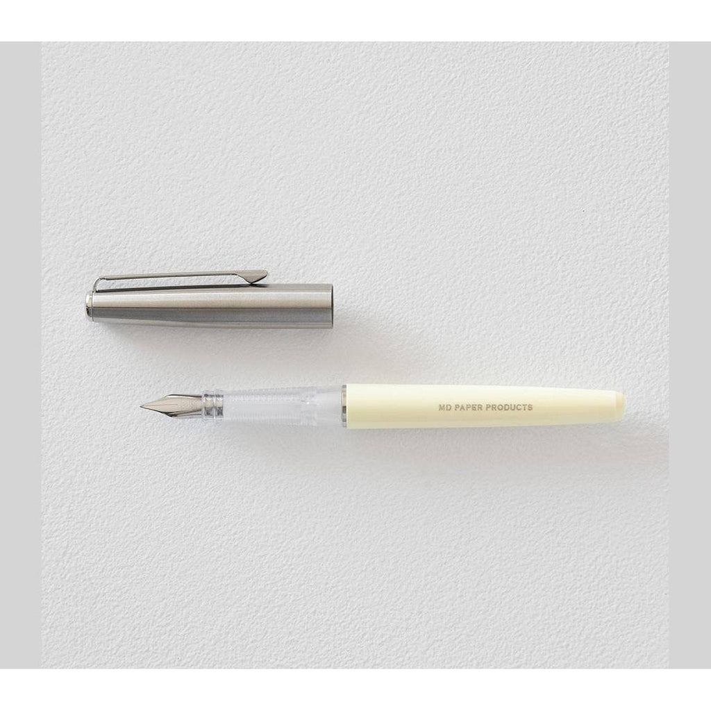 Midori fountain pen with transparent barrel and cream grip lying next to silver cap on white background